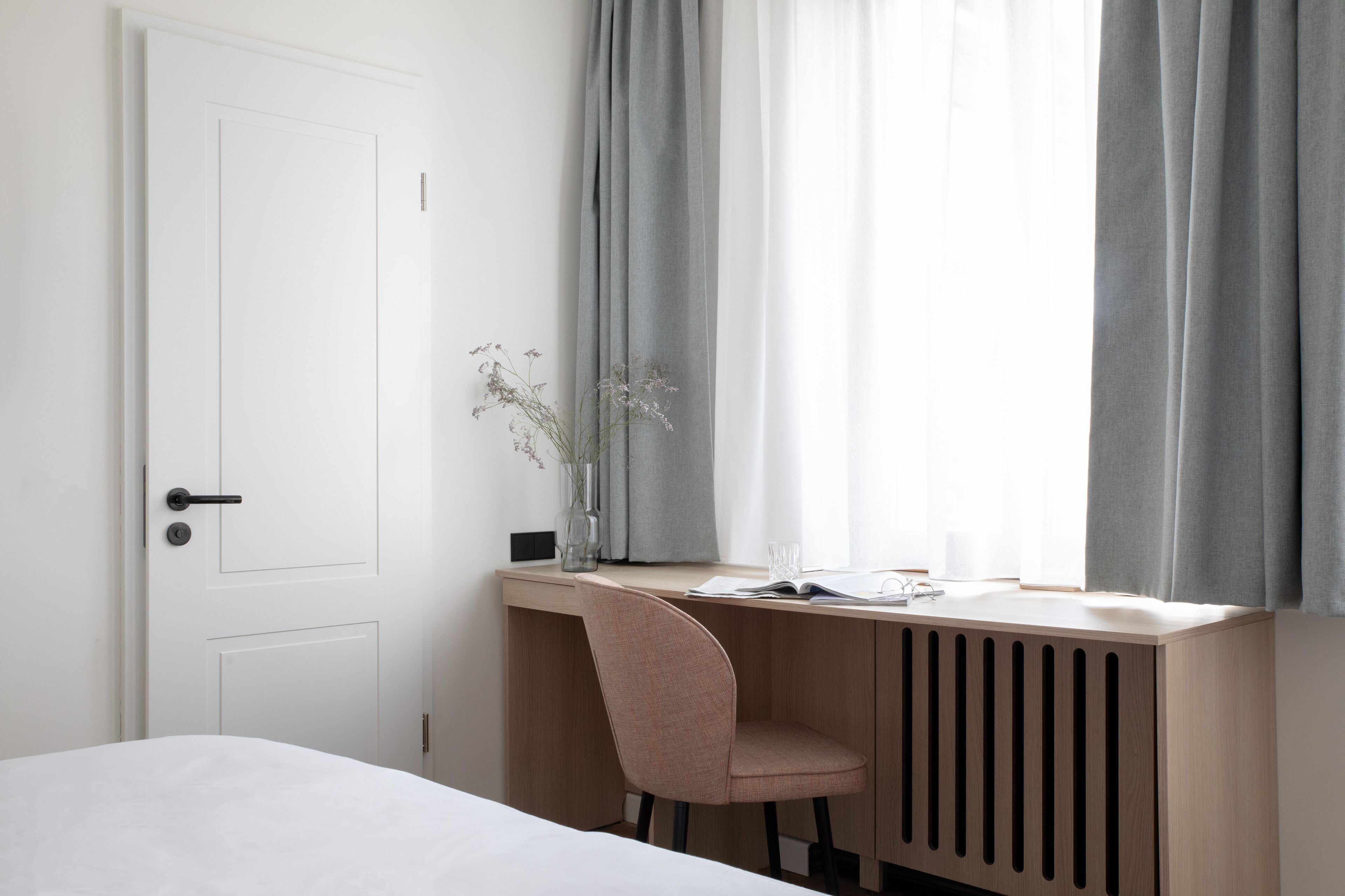 Una stanza minimalista con un letto, una scrivania con una sedia, un termosifone e una finestra con tende.