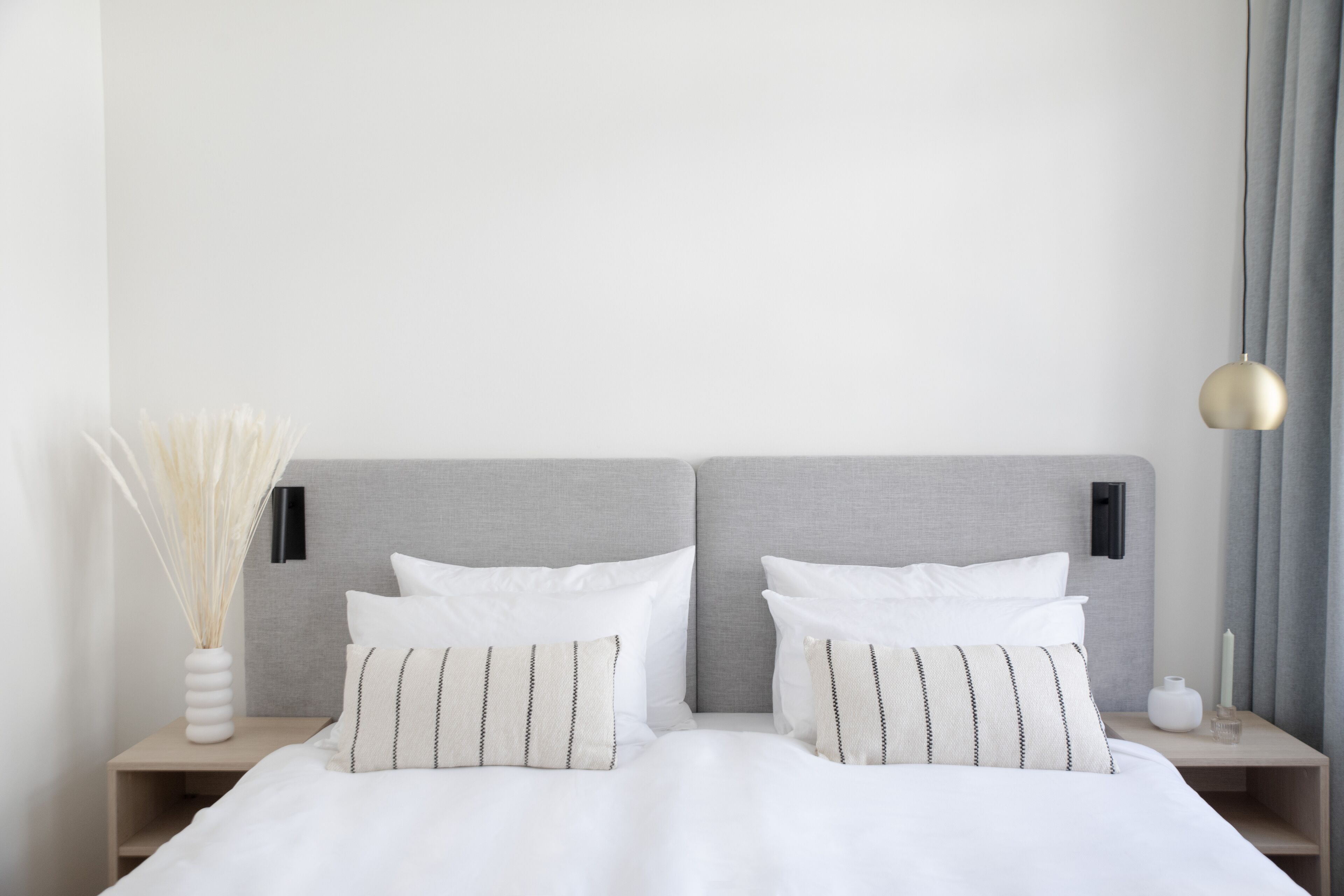 A neatly made bed with white bedding and striped pillows, a bedside table with a vase of dried plants, and a modern pendant light.