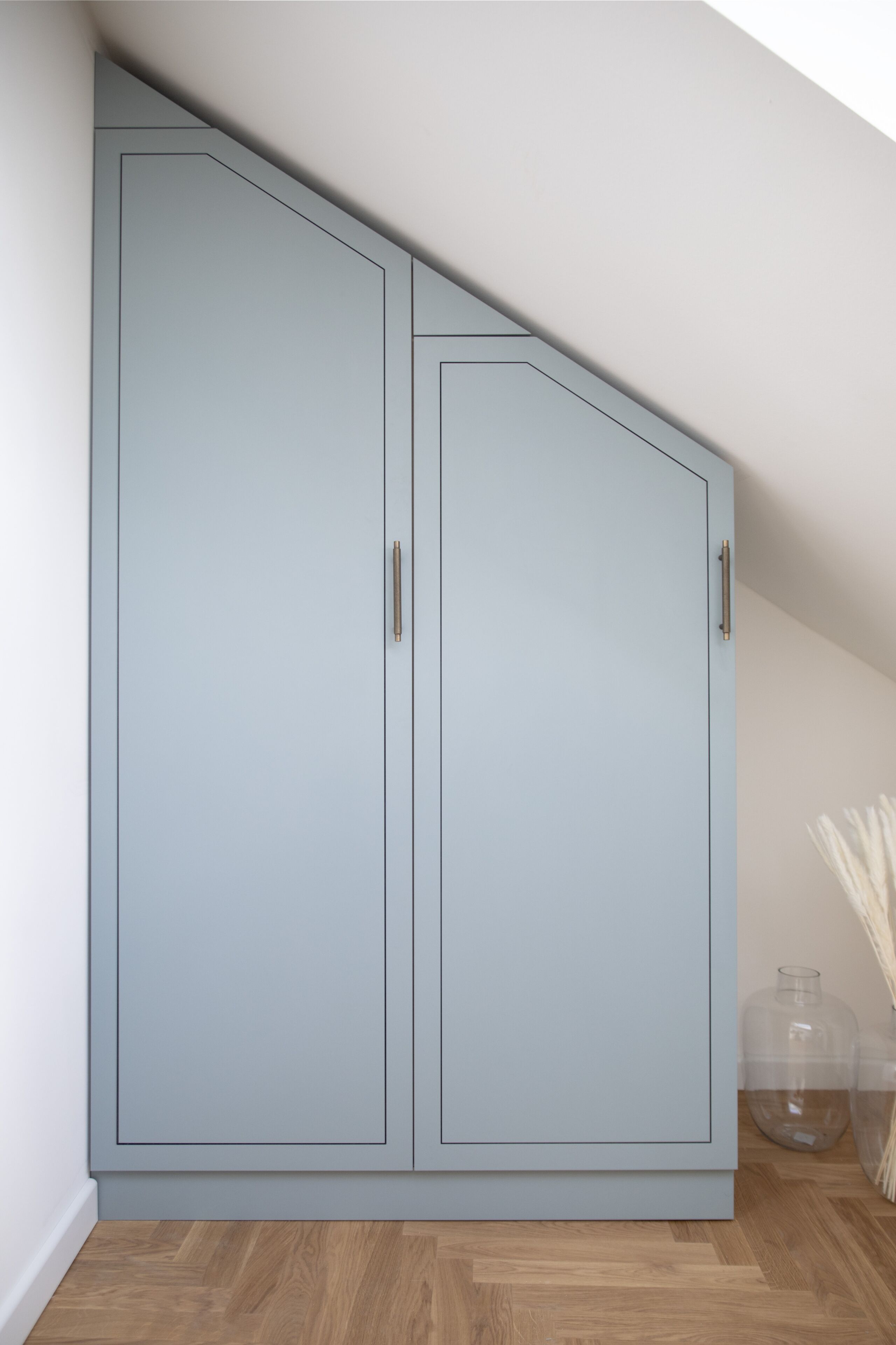 Quarto com guarda-roupa azul claro, piso de madeira e um vaso de vidro com flores brancas.