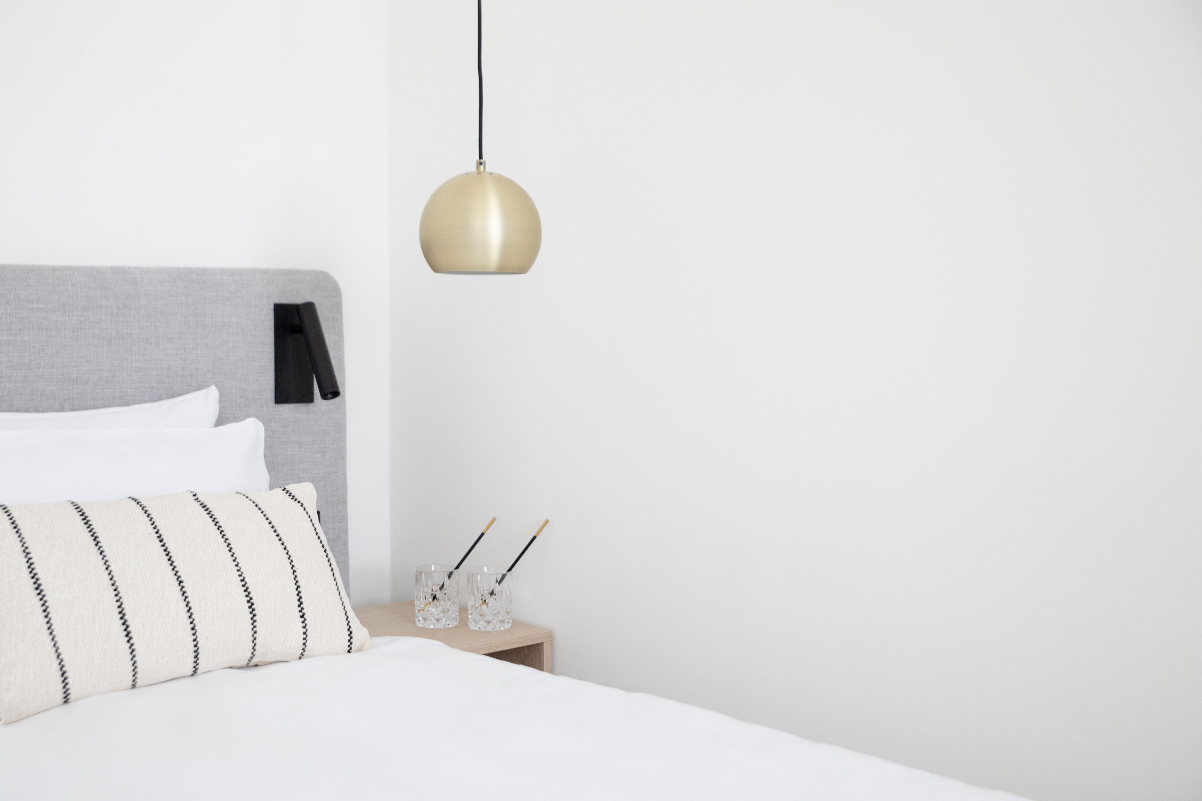 A neatly made bed with a striped pillow, a bedside table with two glasses, and a hanging pendant light.