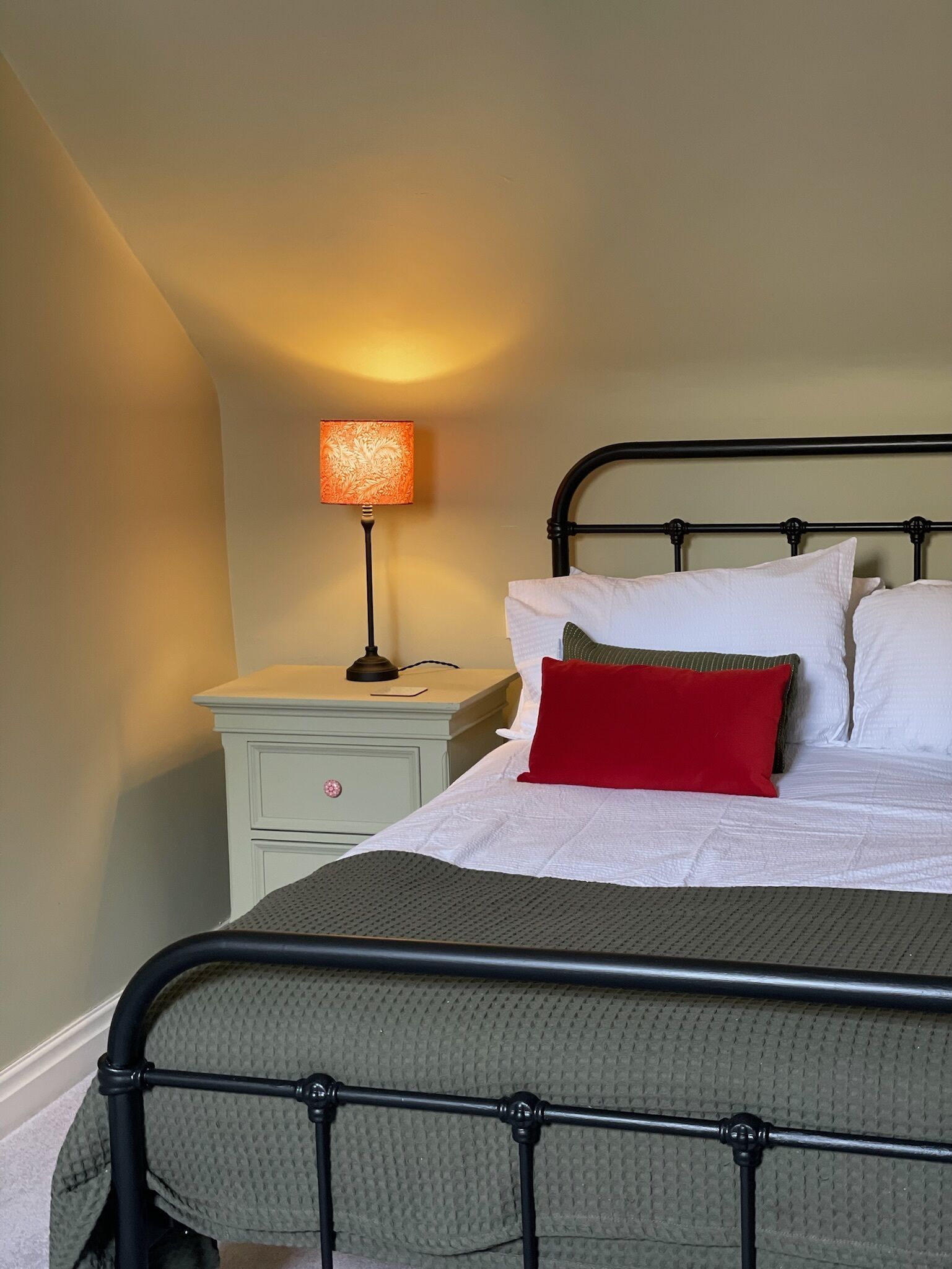 A neatly made bed with a black metal frame, white bedding, and a red pillow. There is a small bedside table with a lamp and a nightstand with a drawer.