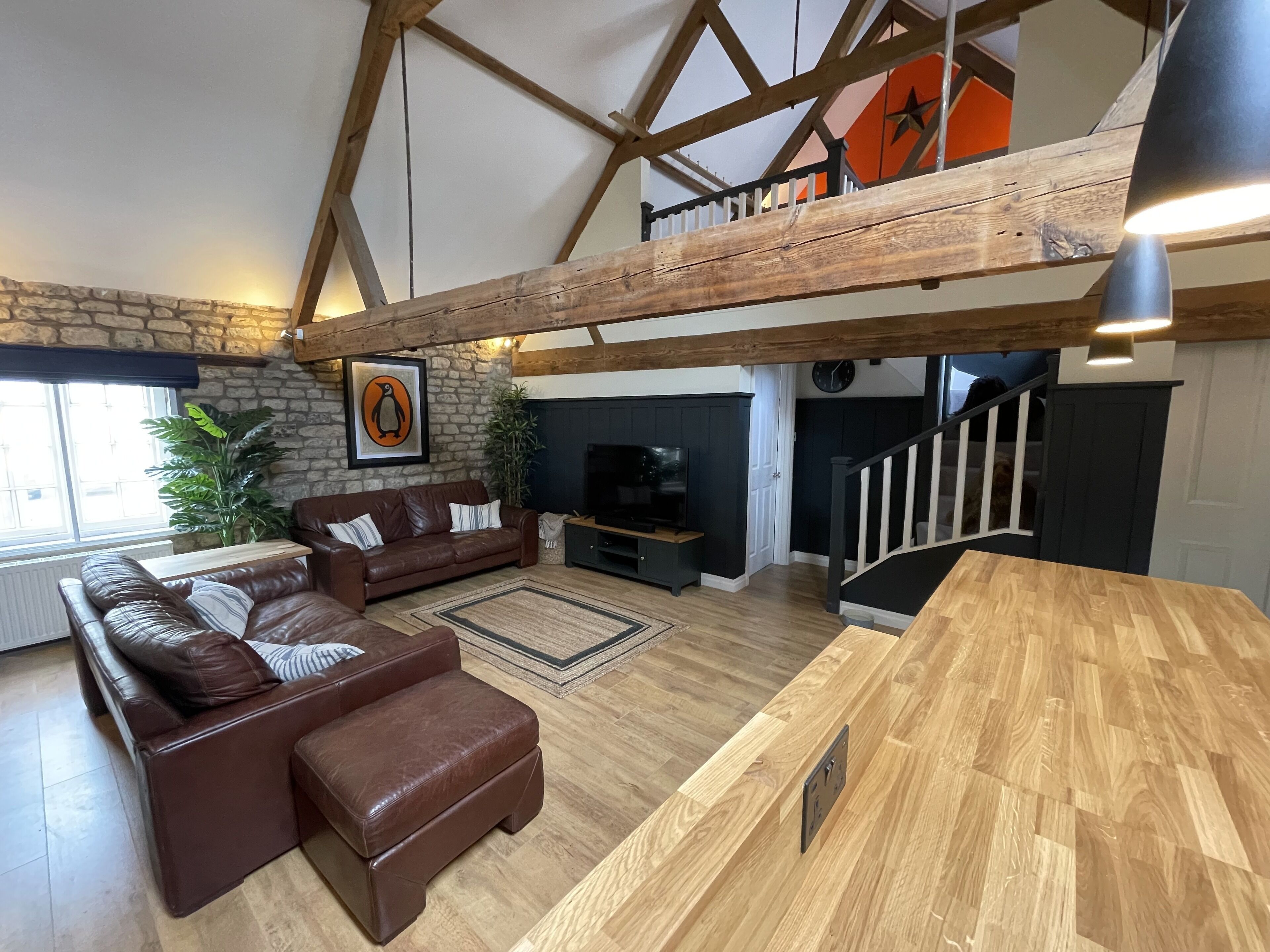 A spacious living area with a stone wall, wooden beams, a brown leather sofa, a wooden coffee table, and a staircase leading to an upper level.