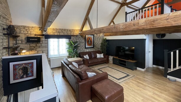 A spacious living area with a stone wall, wooden beams, a brown leather sofa, a television, and a fireplace.