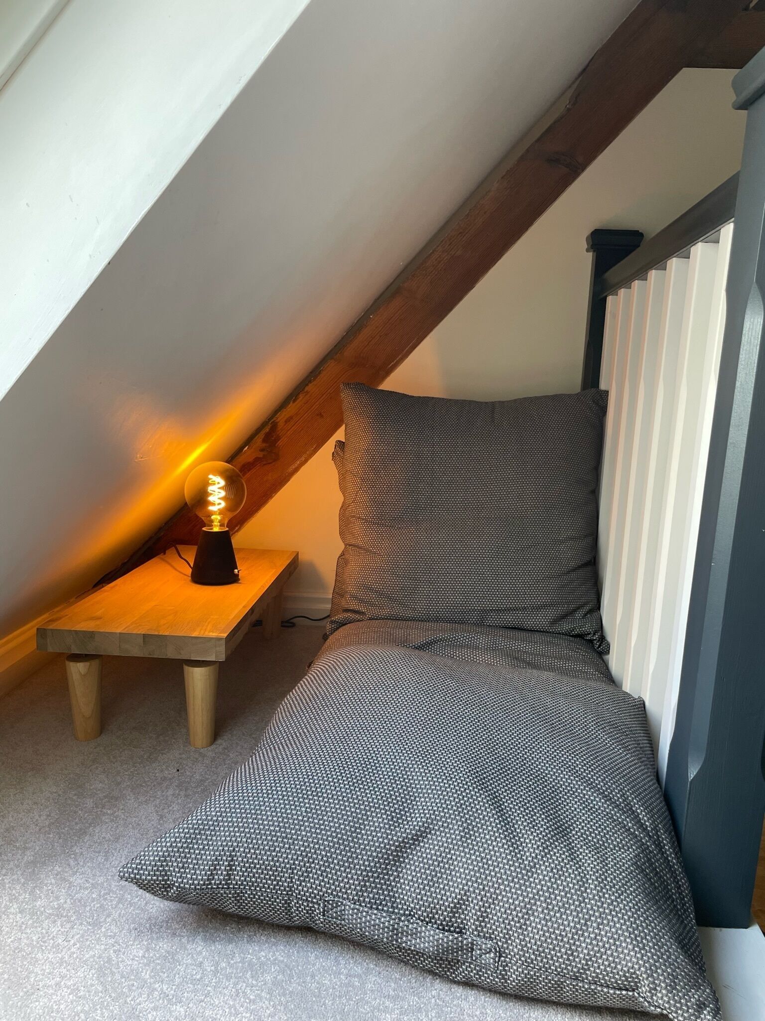 Un dormitorio acogedor en el desván, con techo inclinado, una mesita de noche de madera, una lámpara y una colcha estampada.