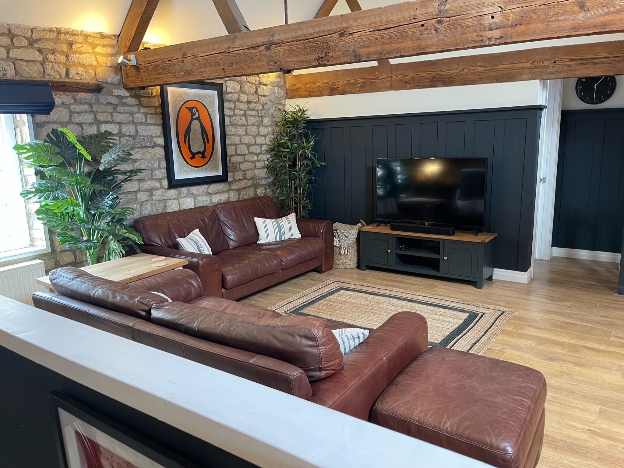 A living room with a brown leather sofa, a flat-screen TV, wooden beams, stone walls, and large green plants.