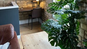 A cozy room with a stone wall, wooden beams, a desk, a chair, a table, a couch, and large green plants.