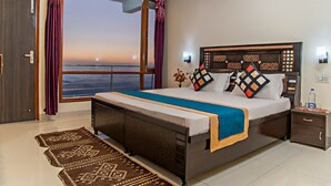 A bedroom with a large bed, a nightstand, a window with curtains, and a view of the sea. - The Bryll Homes (Krishangarh)
