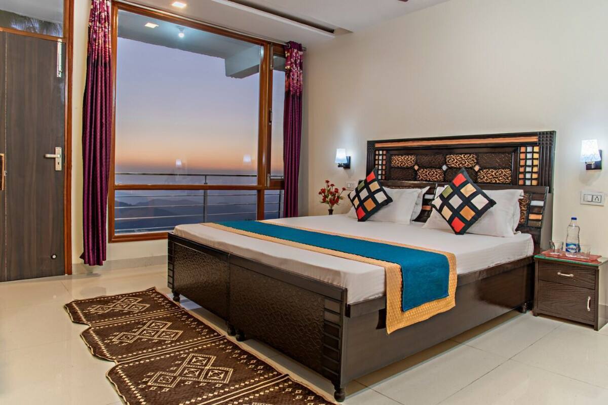 A bedroom with a large bed, a nightstand, a window with curtains, and a view of the sea.