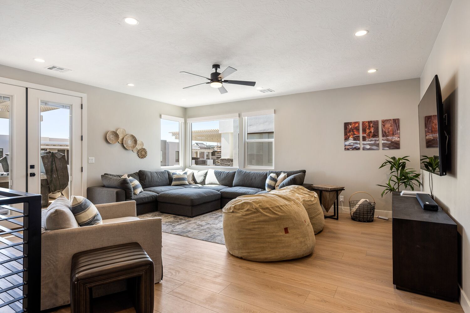 A modern living room with a sectional sofa, a coffee table, and a bean bag. There is a ceiling fan, a TV mounted on the wall, and a small table with a plant.