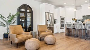 A modern kitchen with white cabinets, a stainless steel refrigerator, and a built-in oven. There are wicker chairs and a leather sofa in the living area. - The Jolly Roger: Desert Color Lagoon + Beaches (St. George)