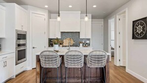 A modern kitchen with white cabinetry, a central island with bar stools, stainless steel appliances, and pendant lighting. - The Jolly Roger: Desert Color Lagoon + Beaches (St. George)
