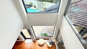 Et rum med højt til loftet, ovenlys, trægulv, en lille rund måtte, en stol og udsigt til en baghave med pool og beplantning.
