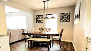 A dining area with a wooden table, chairs, and a bench. There is a window with blinds, a ceiling light fixture, and artwork on the walls. - Luxury 4 Bed Room Near Stadium (Kansas City)