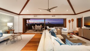 A spacious living room with a large sofa, wooden coffee table, and a ceiling fan. There is a view of the sunset through large windows.