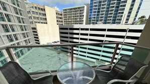 Un balcone con un tavolo in vetro e due sedie in vimini, con vista su un parcheggio e sugli edifici circostanti.