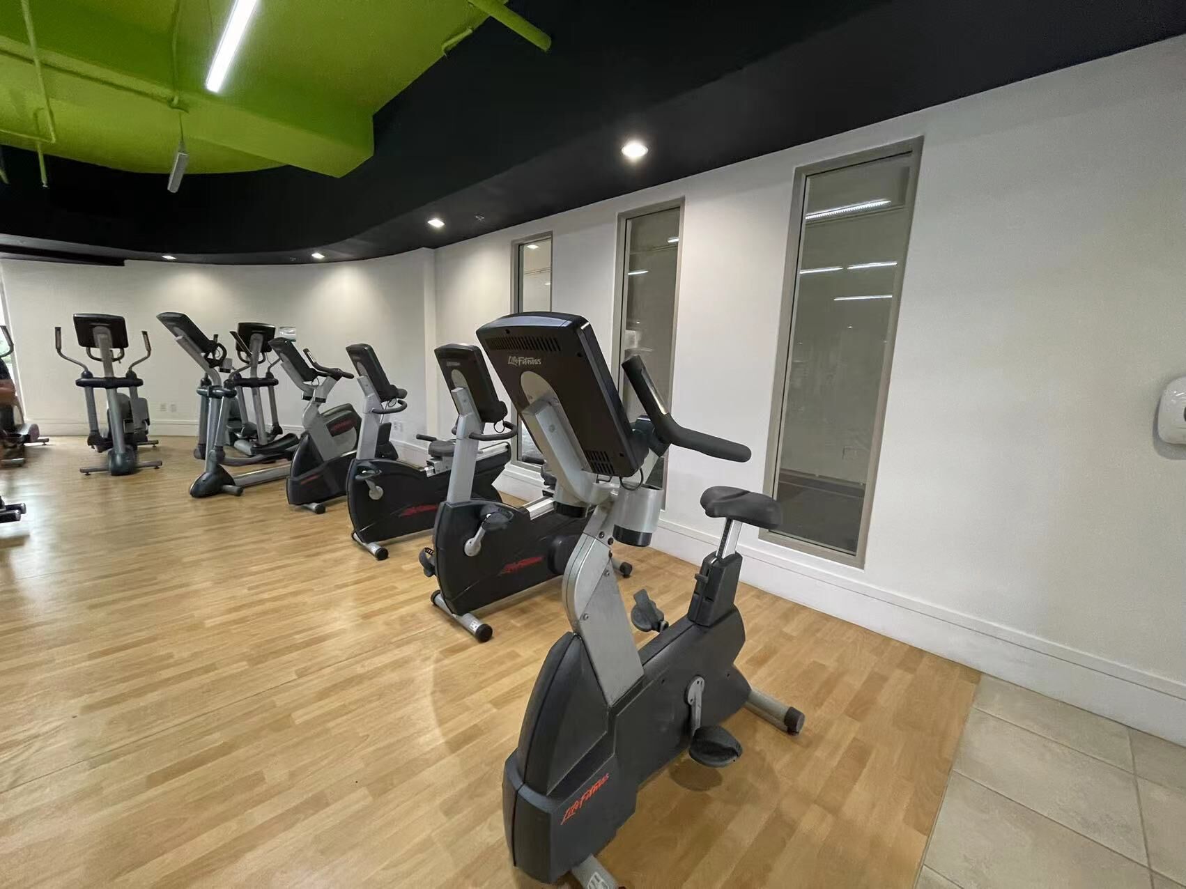 Ett gym med flera konditionsträningsmaskiner, trägolv och stora fönster.