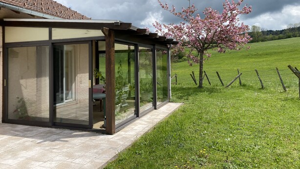A covered outdoor area with a glass enclosure, a wooden pergola, and a view of a green field and a tree with pink blossoms.