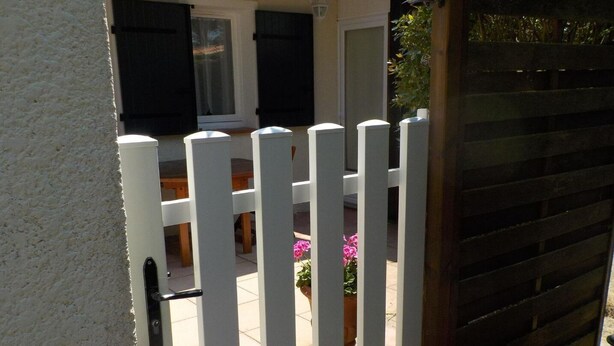 A white gate with vertical bars, a wooden fence, and a patio area with a table and chairs. - Maison Tout Confort à Lacanau Océan (Lacanau)