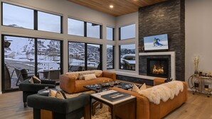 Un salón amplio con un ventanal que ofrece vistas a un paisaje montañoso nevado, una chimenea y un televisor montado encima.