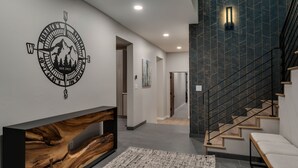 Un interior moderno con una consola de madera, un adorno de pared con forma de brújula, una escalera con barandillas negras y un pasillo que conduce a otra habitación.