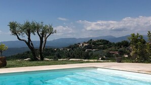 A swimming pool with clear blue water, surrounded by a tiled deck. In the background, there are olive trees and a view of a hilly landscape with buildings. - Villa Provençale Tout Confort Avec vue (Tanneron)