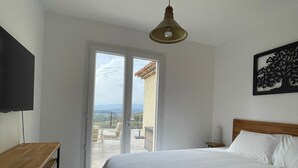 A bedroom with a bed, a wooden table, a TV, a ceiling light, and a view through a glass door to an outdoor terrace. - Villa Provençale Tout Confort Avec vue (Tanneron)