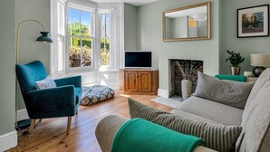 Ein Wohnzimmer mit einem salbeingrünem Sessel, einem beigefarbenen Sofa, einer Holzkabine, einem Fernseher und einem Kamin.