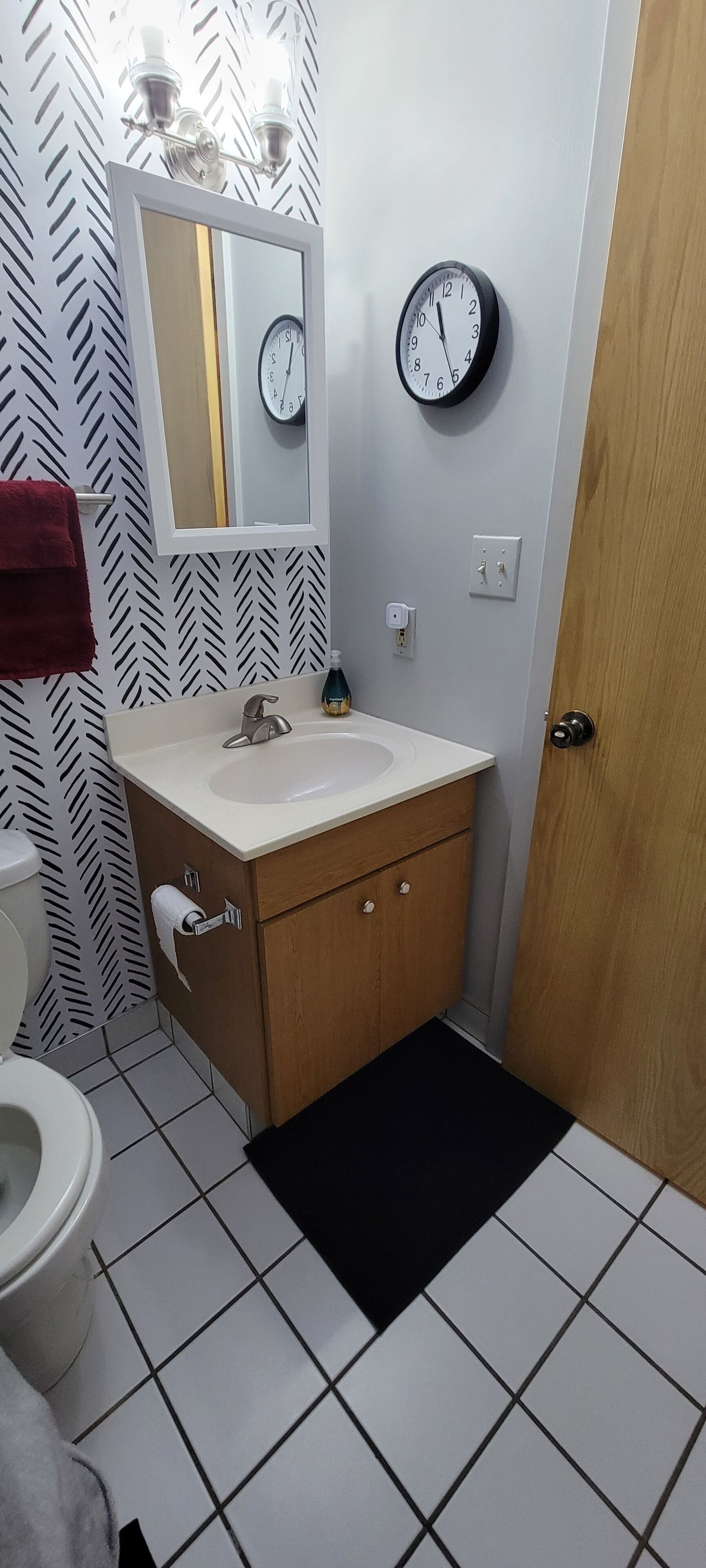 Une salle de bain avec un lavabo blanc, un meuble en bois et une horloge noire accrochée au mur.