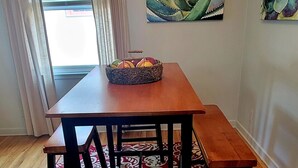 Un comedor con una mesa de madera y bancos, una alfombra estampada y dos cuadros en la pared.