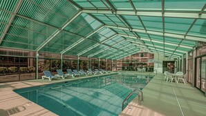En inomhuspool med väggar av genomskinligt glas, solstolar och bord.