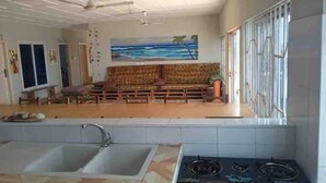 Un salon spacieux comprenant un ensemble de canapés, un tableau représentant une scène de plage et un coin cuisine avec une cuisinière à gaz et un évier.