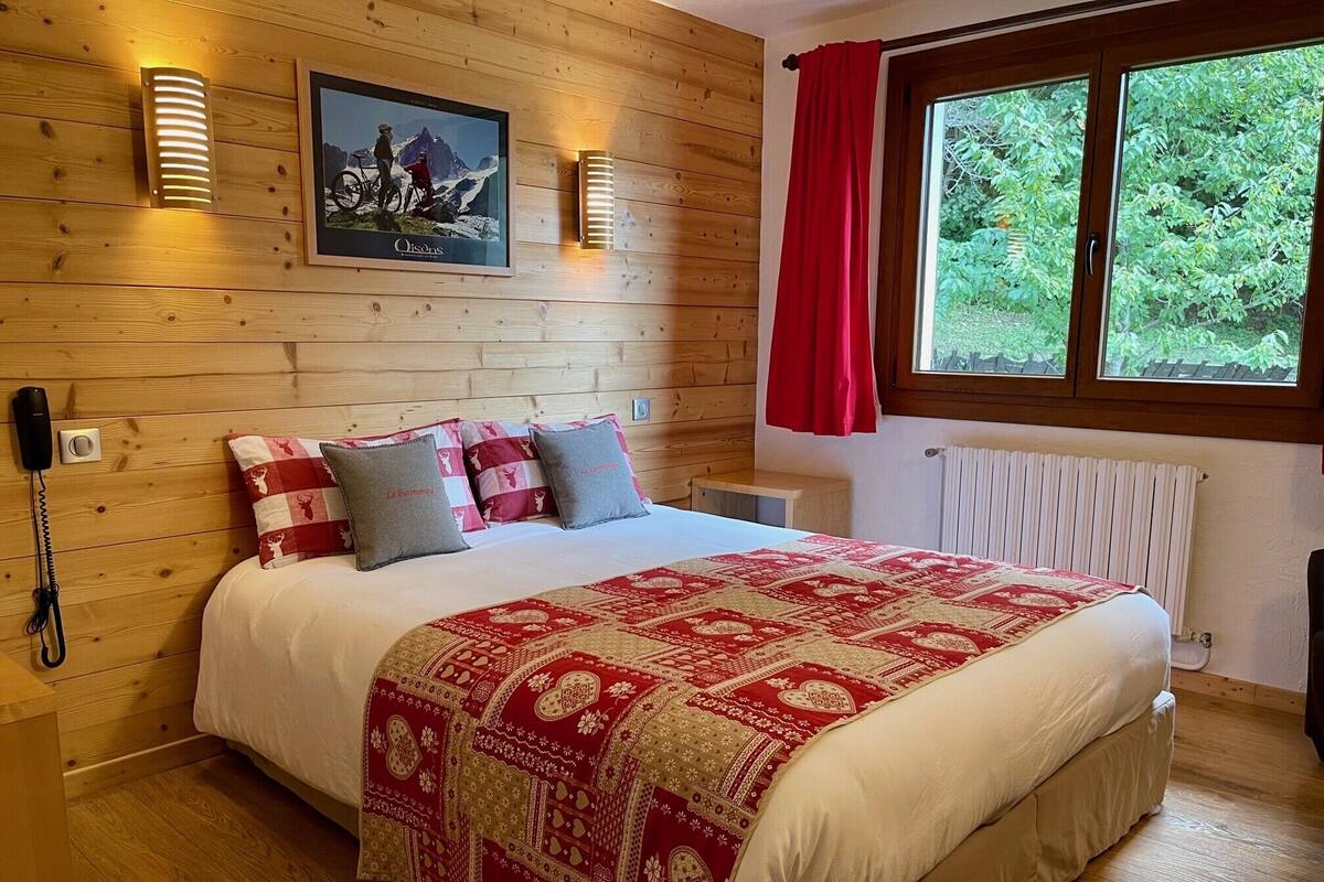 Ein Schlafzimmer mit Holzwänden, einem Bett, Kissen, einem aufgehängten Bild, einem Telefon und einem Fenster mit Blick auf Grünflächen.