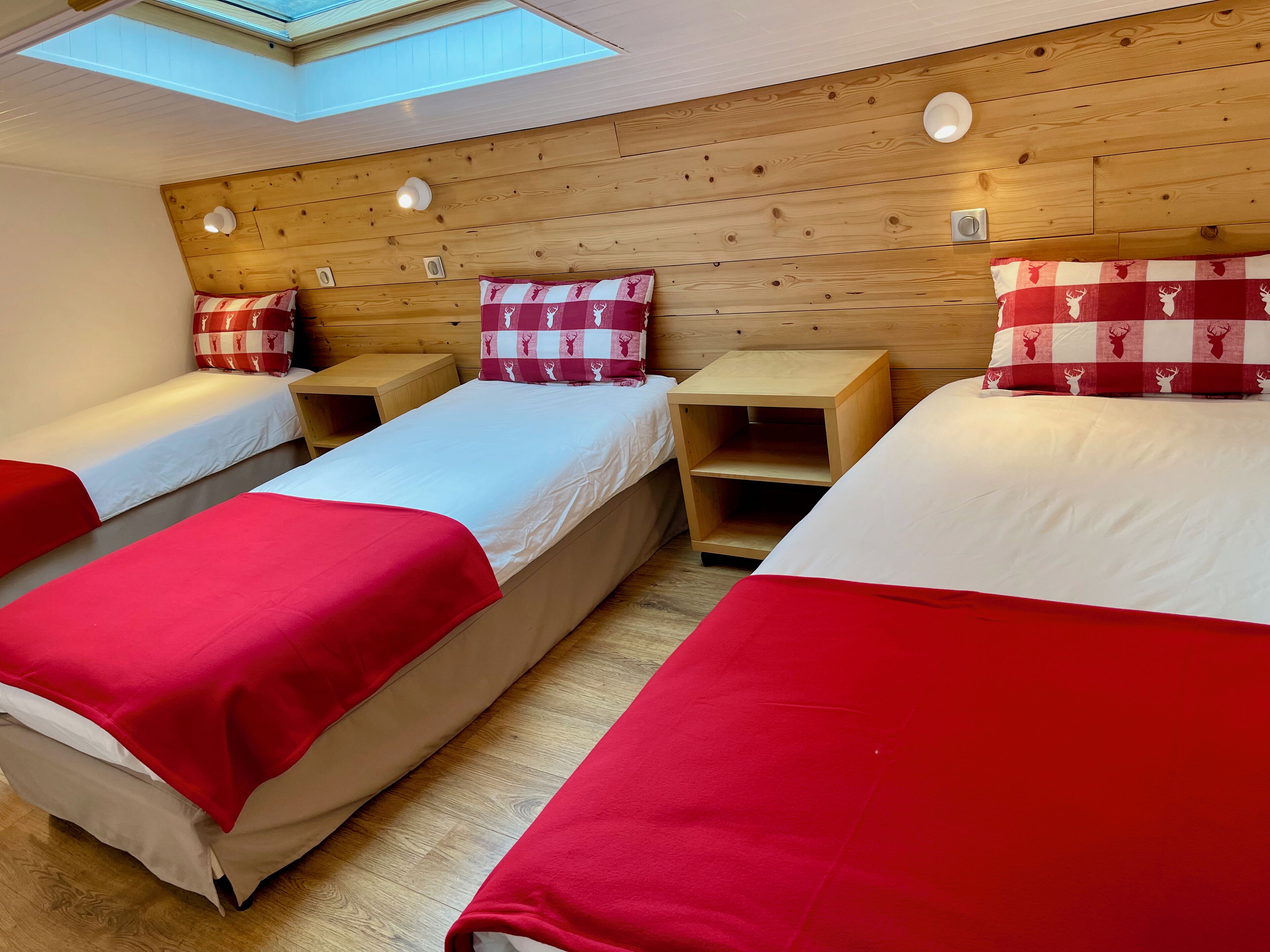A room with three beds, each with a red blanket and matching pillows, wooden headboards, and a skylight above.