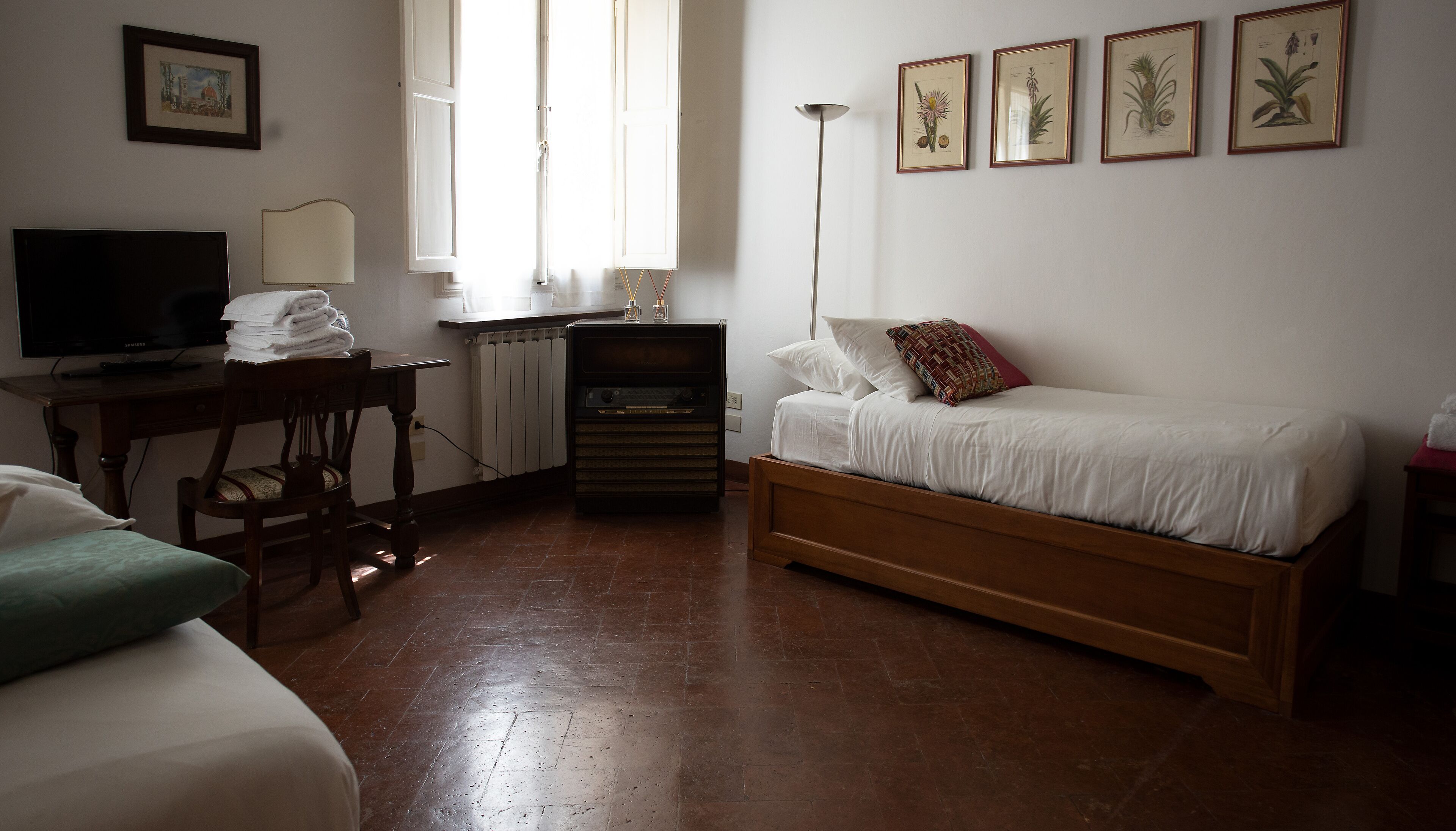 A bedroom with a bed, a desk with a computer, a chair, a radiator, and framed botanical prints on the wall.