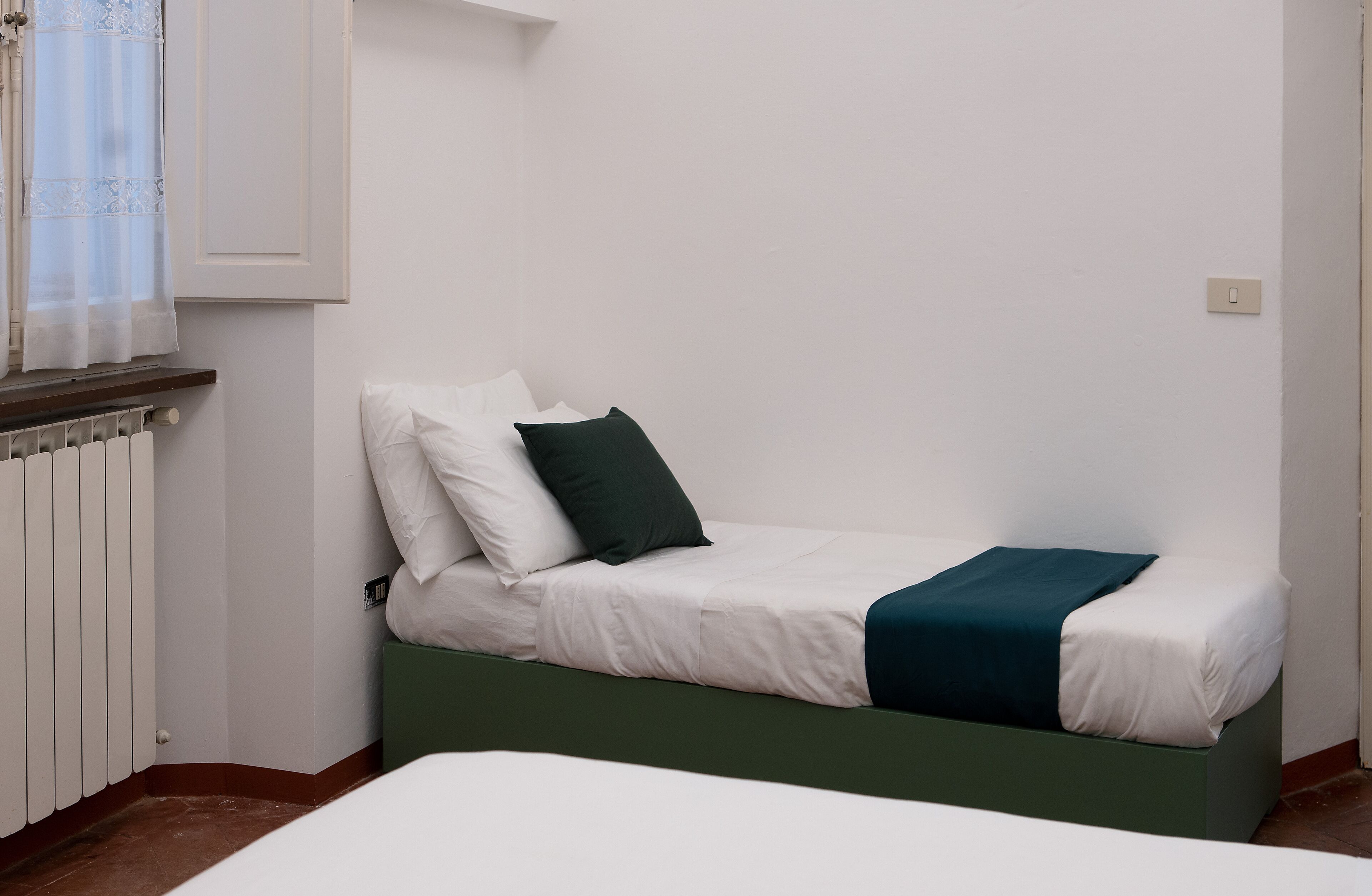 A single bed with white bedding and a dark green throw, a window with sheer curtains, and a white radiator.
