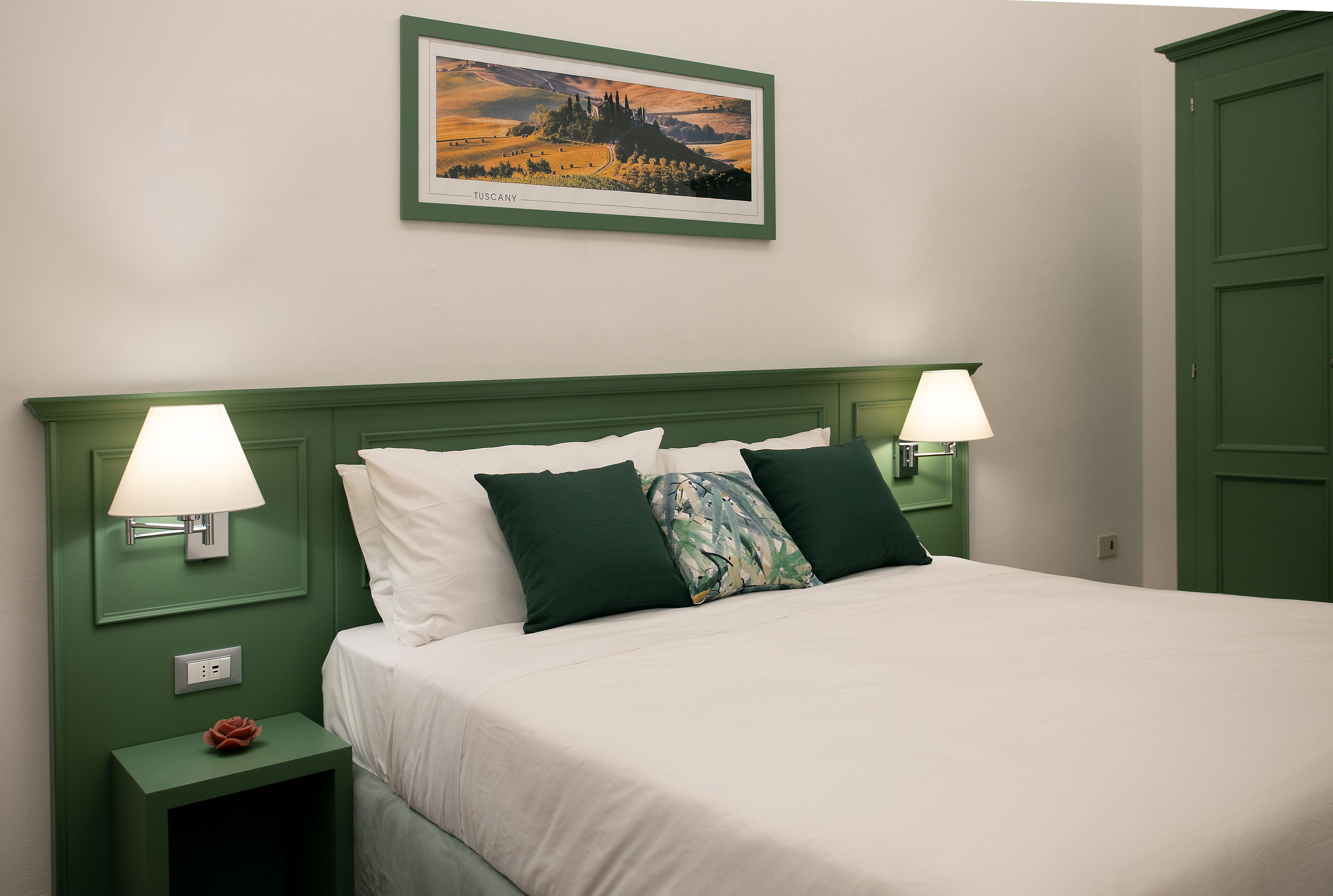 A neatly made bed with white linens and green pillows, flanked by two bedside lamps on matching green nightstands. A framed picture of Tuscany hangs above the bed.