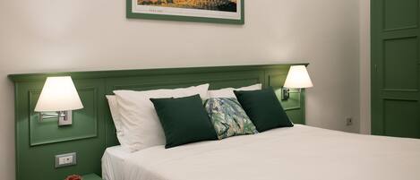 A neatly made bed with white linens and green pillows, flanked by two bedside lamps on matching green nightstands. A framed picture of Tuscany hangs above the bed.