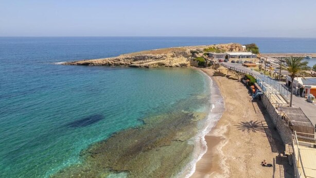 Un hotel frente al mar con vista a la playa, un promontorio rocoso y un mar azul cristalino.