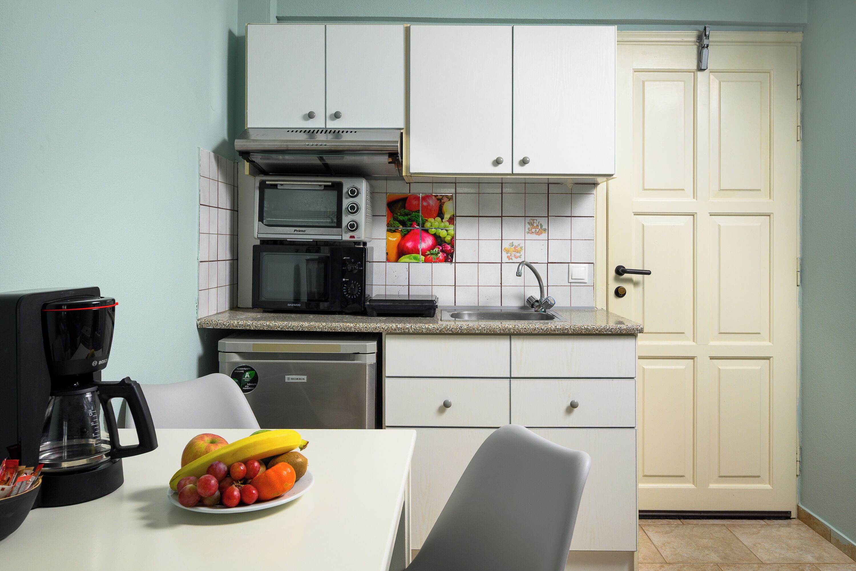 A compact kitchen with white cabinets, a microwave, a coffee maker, and a fruit bowl on the counter.