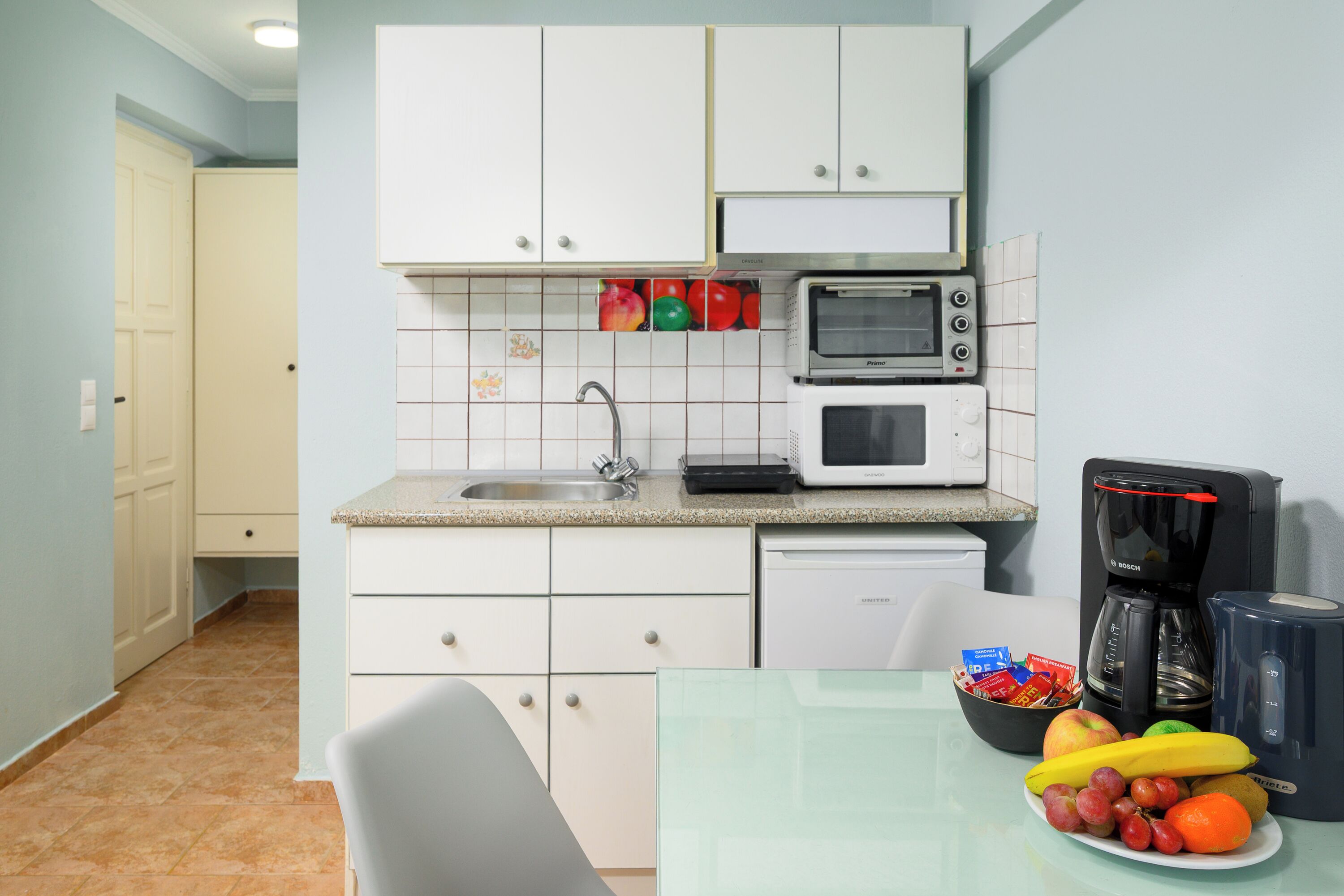 Une cuisine compacte avec des meubles blancs, un four à micro-ondes, une machine à café et un panier à fruits posé sur le comptoir.