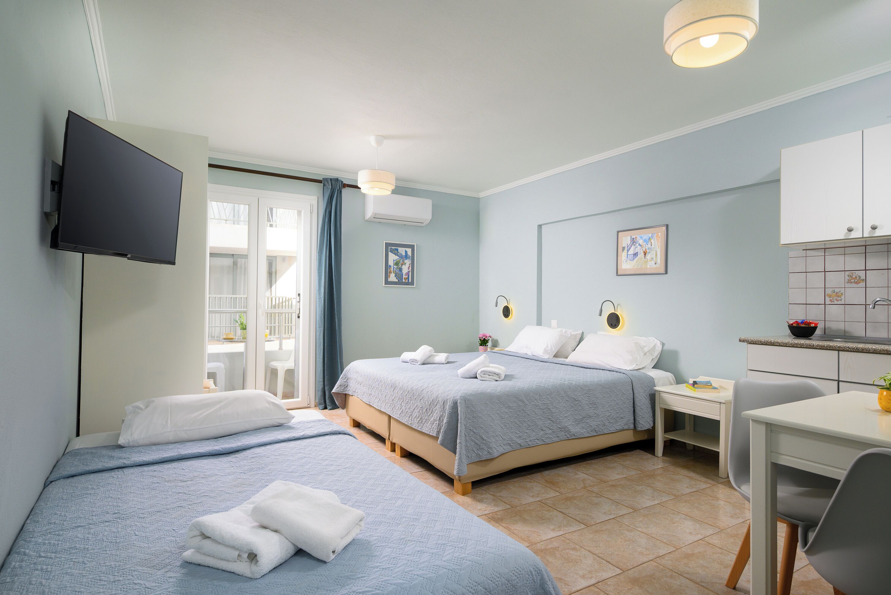 A hotel room with two beds, a TV, a desk, and a kitchenette.