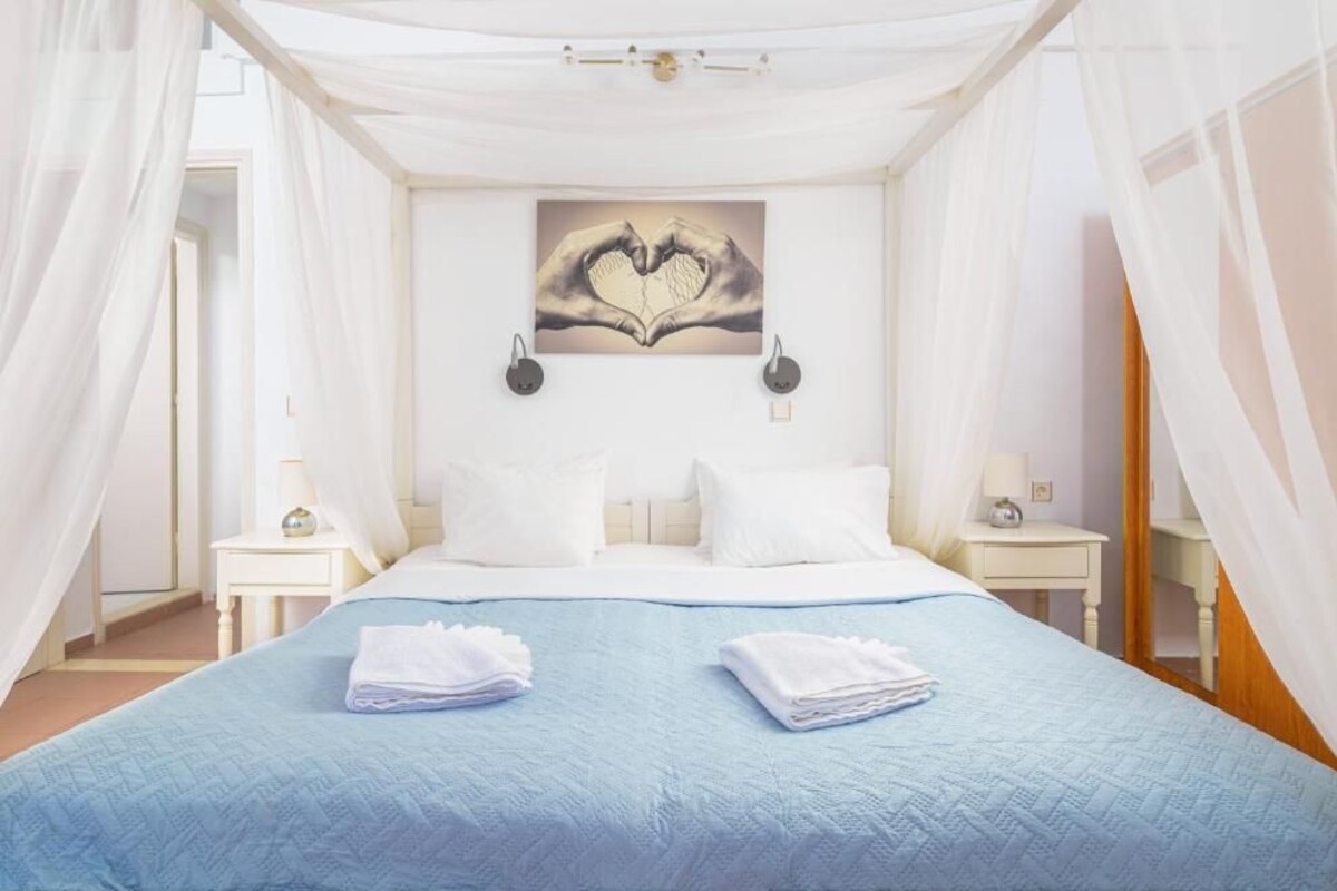 A bedroom with a bed, white curtains, a wall-mounted heart-shaped artwork, and two bedside tables with lamps.