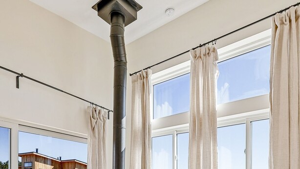 Een moderne woonkamer met een houtkachel, grote ramen en uitzicht op een huis en landschap.