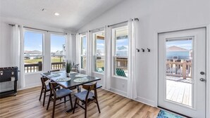Un comedor amplio con mesa y sillas de madera, ventanas grandes con cortinas y vistas a una terraza y montañas.