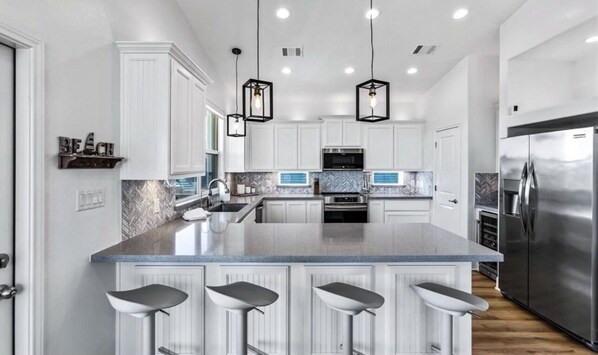 Une cuisine moderne avec des meubles blancs, un grand îlot central doté de tabourets, des appareils en acier inoxydable et un éclairage suspendu.