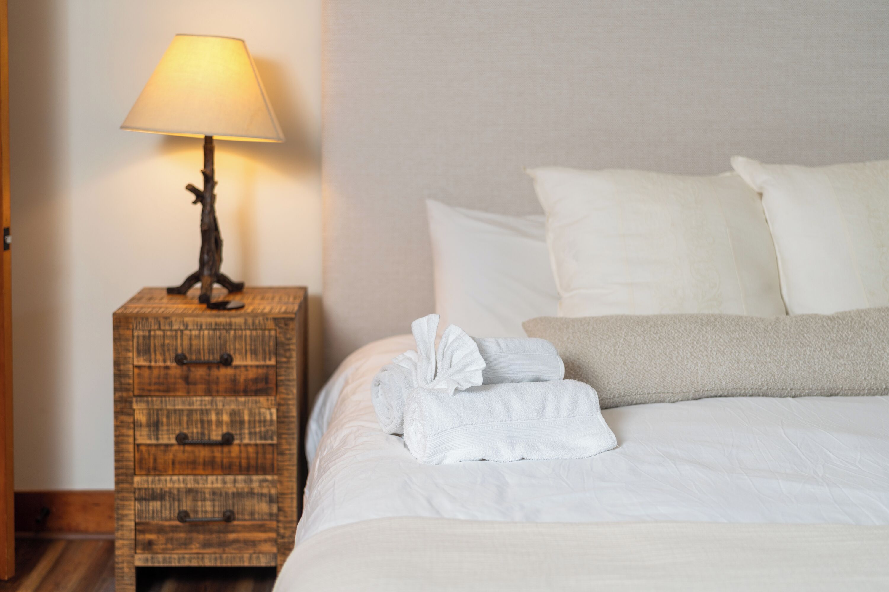 A neatly made bed with white linens and a folded towel, a wooden nightstand with a lamp, and a plain wall in the background.
