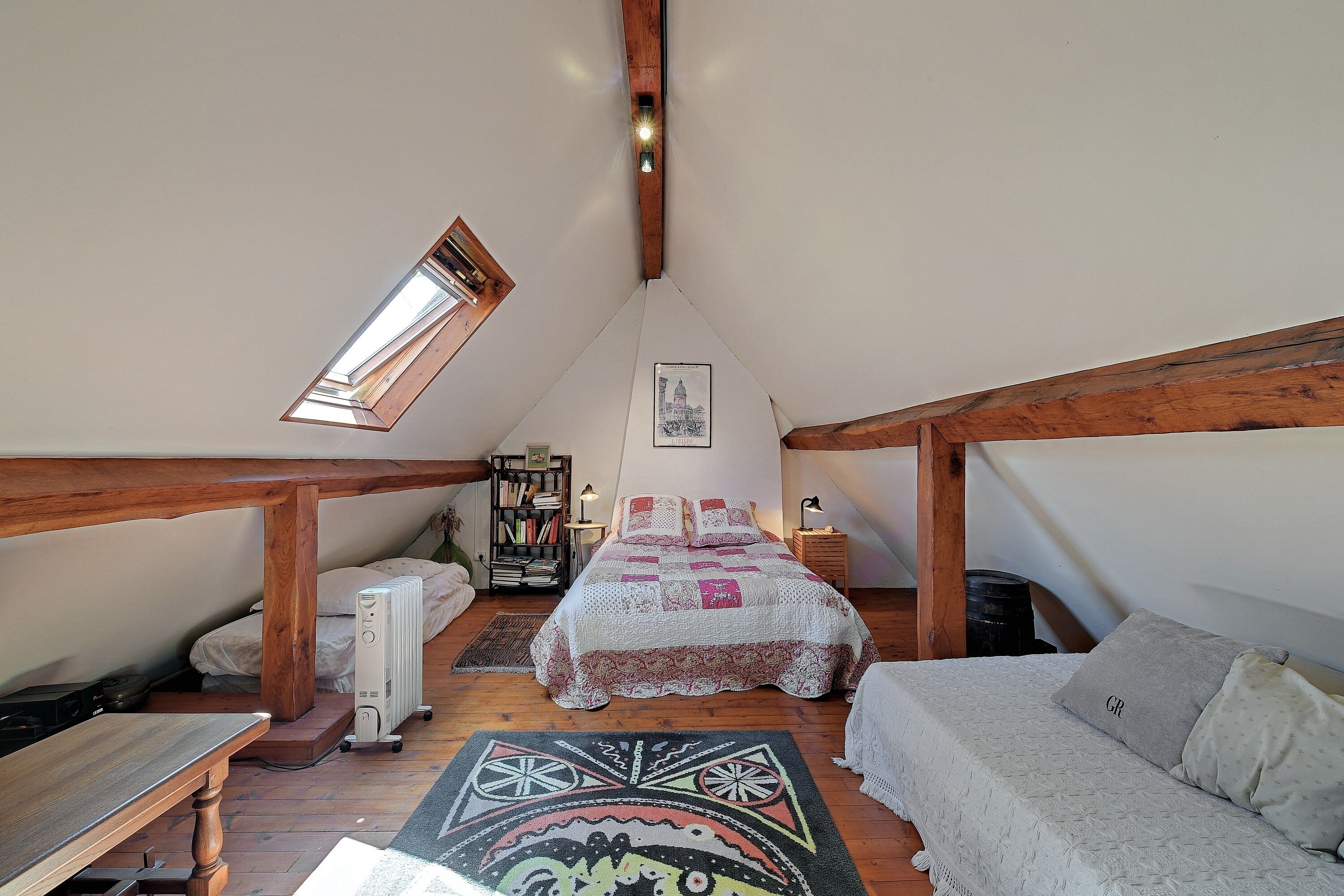 A cozy attic bedroom with a sloped ceiling, a skylight, a bed with a patterned quilt, a small table, and a bookshelf.