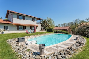 A two-story house with a swimming pool, surrounded by a stone border and a grassy lawn.