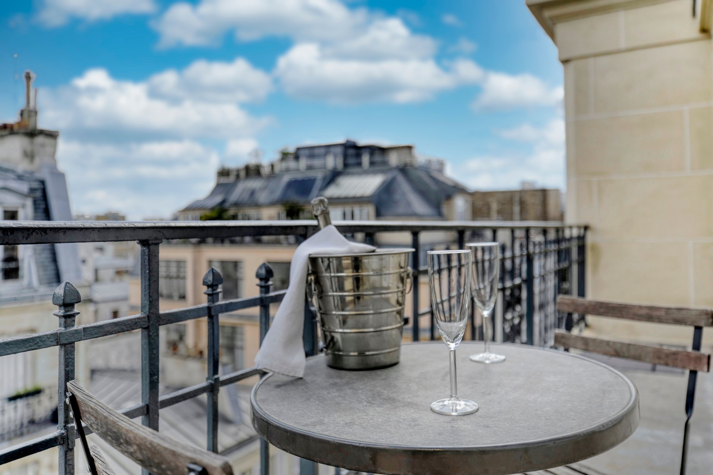A round metal table with two wine glasses and a metal bucket on a balcony with a metal railing.
