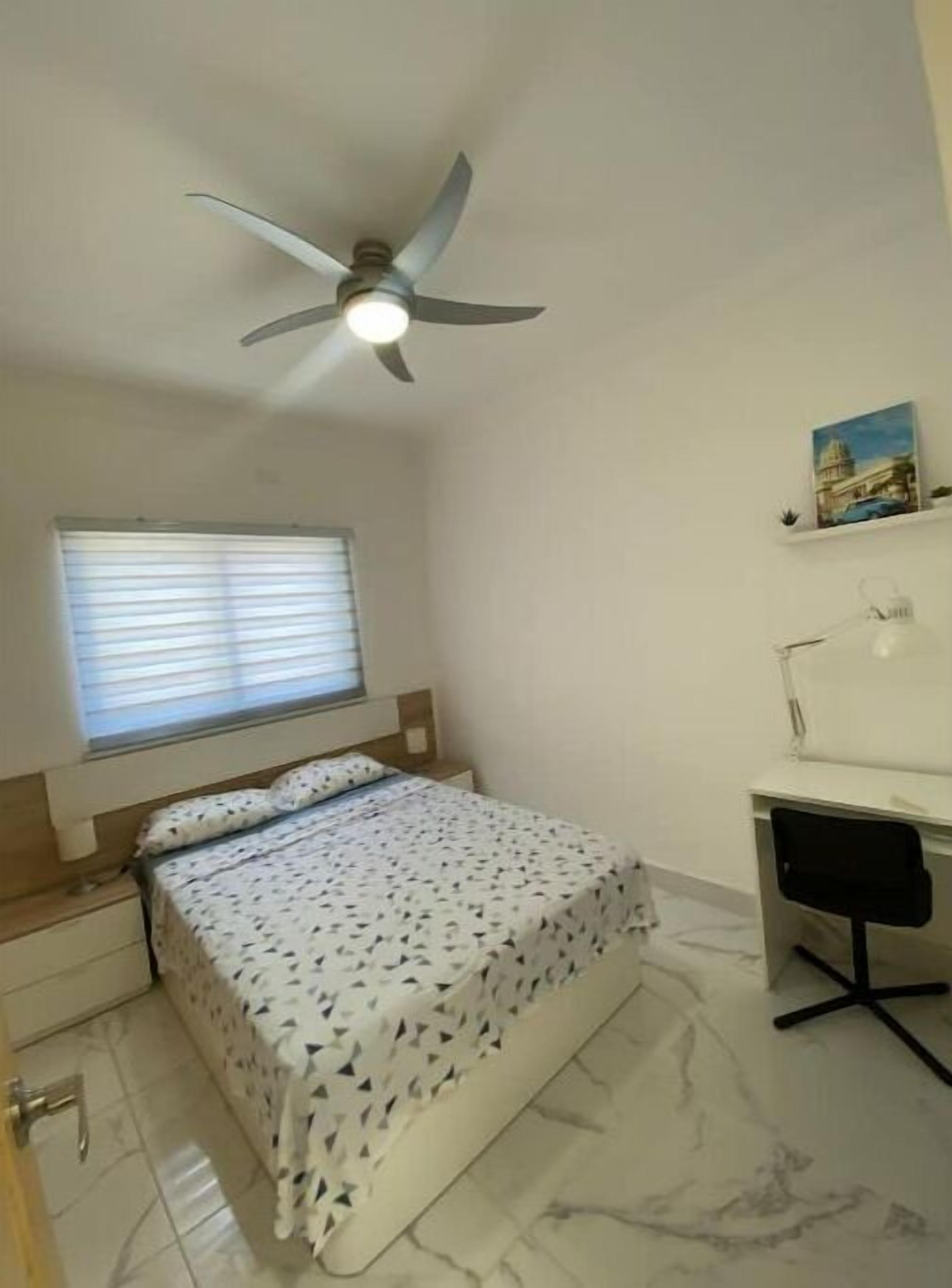 A bedroom with a bed, a desk, a chair, a ceiling fan, and a window with blinds.